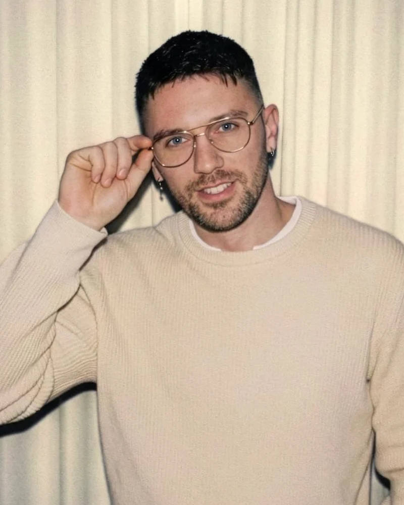 Tomaso Falchi mit Brille im hellen Strickpullover, lächelnd, vor einem Vorhang