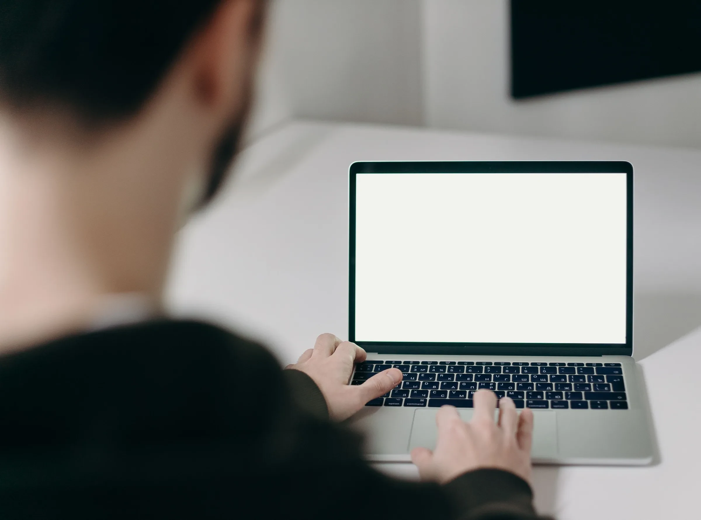 Ein Mann im dunklen Kapuzenpullover sitzt, von hinten gesehen, in einem hellen Raum am Laptop, die Hände auf der Tastatur