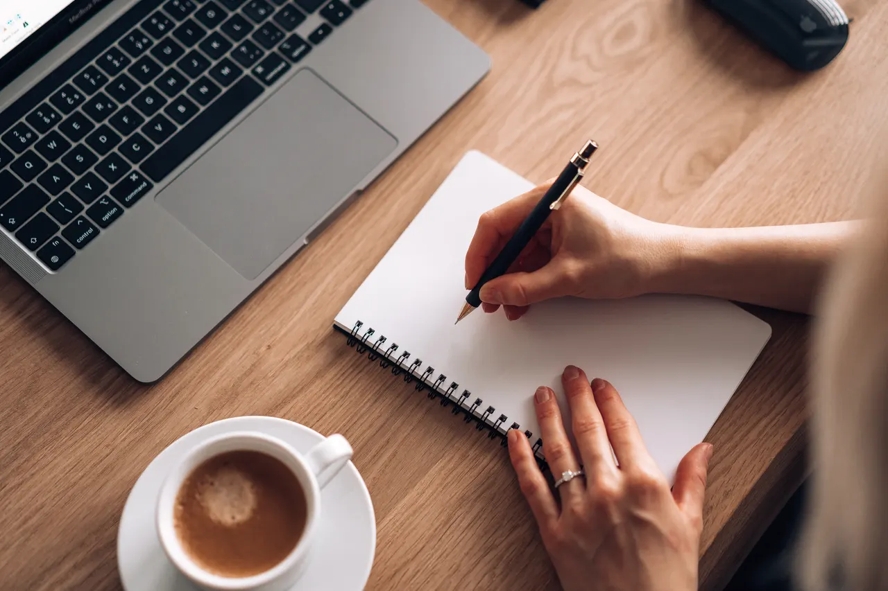 Eine Hand schreibt mit Stift in ein Notizbuch auf einem Holztisch, daneben ein Laptop
